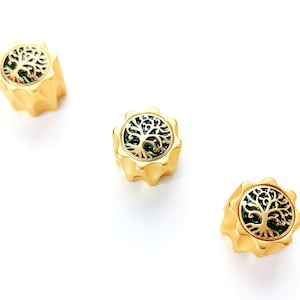 Könnte beinhalten: Drei goldfarbene Perlen mit einem Lebensbaum-Design. Jede Perle hat einen gewellten Rand und einen grünen Hintergrund hinter dem Baumdesign. Die Perlen werden auf weißem Hintergrund dargestellt.