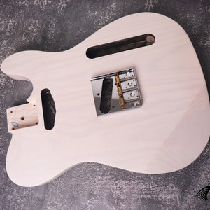 Könnte beinhalten: Unfertiger, hellfarbiger Gitarrenkorpus mit Chrombrücke und Hardware. Der Korpus hat gefräste Abschnitte für Tonabnehmer und einen Hals. Das Bild ist vor einem strukturierten, dunkelgrauen Hintergrund.