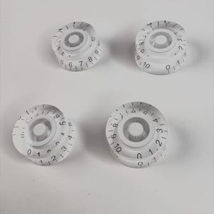 May include: Four white plastic knobs with black numbers and markings. The knobs are for a guitar or other musical instrument.