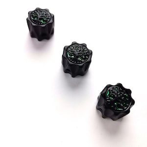 May include: Three black bottle stoppers featuring a green tree of life design. The stoppers have a ridged, star-like shape. The stoppers are set against a white background.