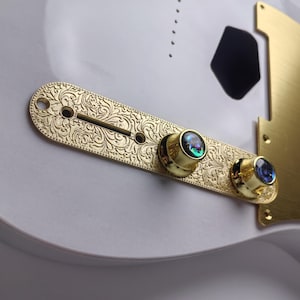 May include: Close-up of a gold-colored guitar control plate with intricate floral engraving. The plate features two control knobs with iridescent inlays. A matching gold pickguard is also visible, set against a white guitar body.