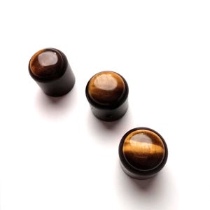 May include: Three brown bottle stoppers with tiger's eye gemstone tops. The stoppers have a dark brown cylindrical base and a polished, rounded gemstone. The gemstones have brown and gold tones.