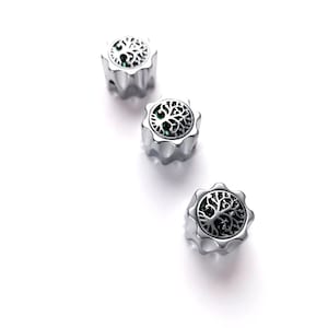 Puede incluir: Tres cuentas plateadas con un diseño de árbol de la vida. Las cuentas tienen una parte superior circular con un patrón de árbol detallado y un borde festoneado. El fondo es blanco.