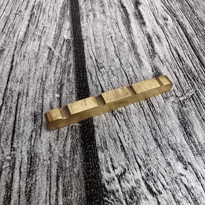 May include: A rectangular, brass-colored guitar nut with three raised sections. The nut is positioned diagonally on a gray and black wood-grain surface, providing a contrast in texture and color.