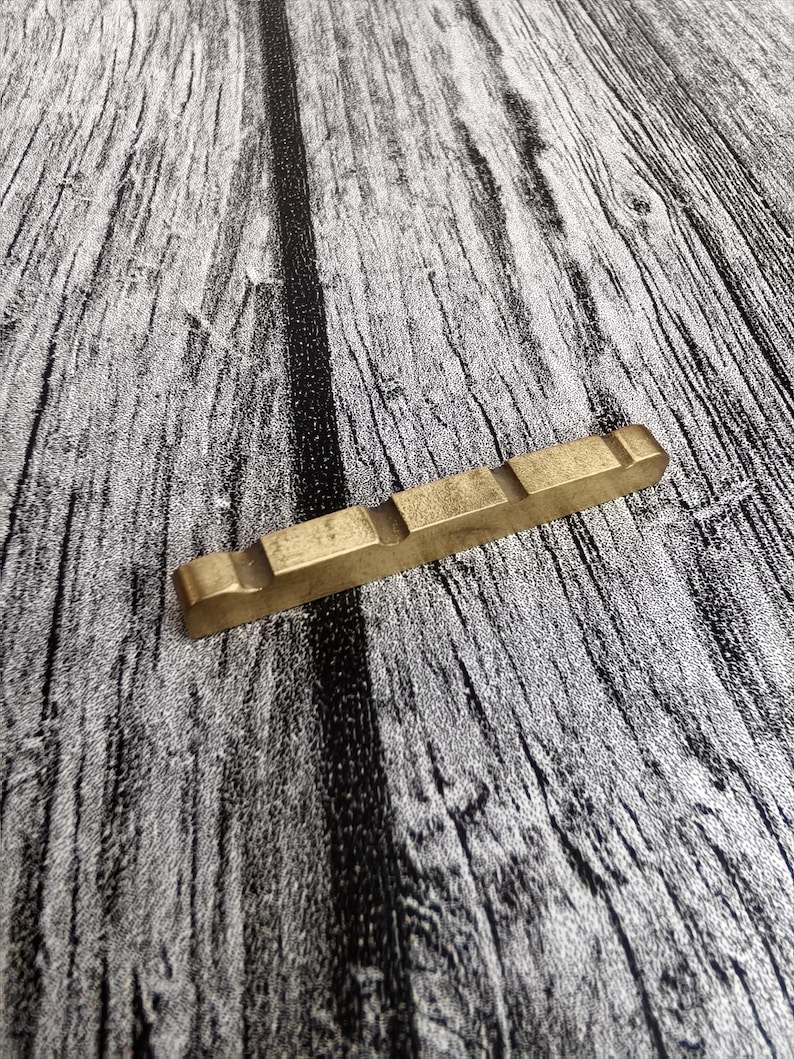 May include: A rectangular, brass-colored guitar nut with three raised sections. The nut is positioned diagonally on a gray and black wood-grain surface, providing a contrast in texture and color.