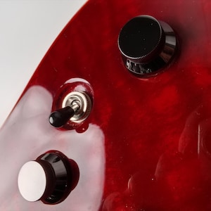 Op de afbeelding: Close-up van een glanzende, rode elektrische gitaarbody met zwarte bedieningsknoppen en een tuimelschakelaar. De afwerking van de gitaar heeft een levendig, wervelend patroon en de bedieningselementen zijn netjes gerangschikt.