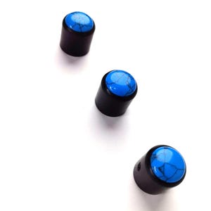 Peut inclure: Trois bouchons cylindriques avec une base marron foncé et un dessus bleu vif marbré. Les bouchons sont disposés en diagonale sur un fond blanc. Les dessus bleus ont un aspect turquoise.