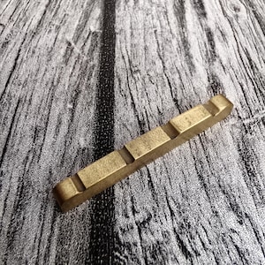 May include: A rectangular, brass-colored guitar nut with four grooves. The nut is made of a solid, metallic material and is designed to fit on a guitar. The background is a gray and white wood grain pattern.