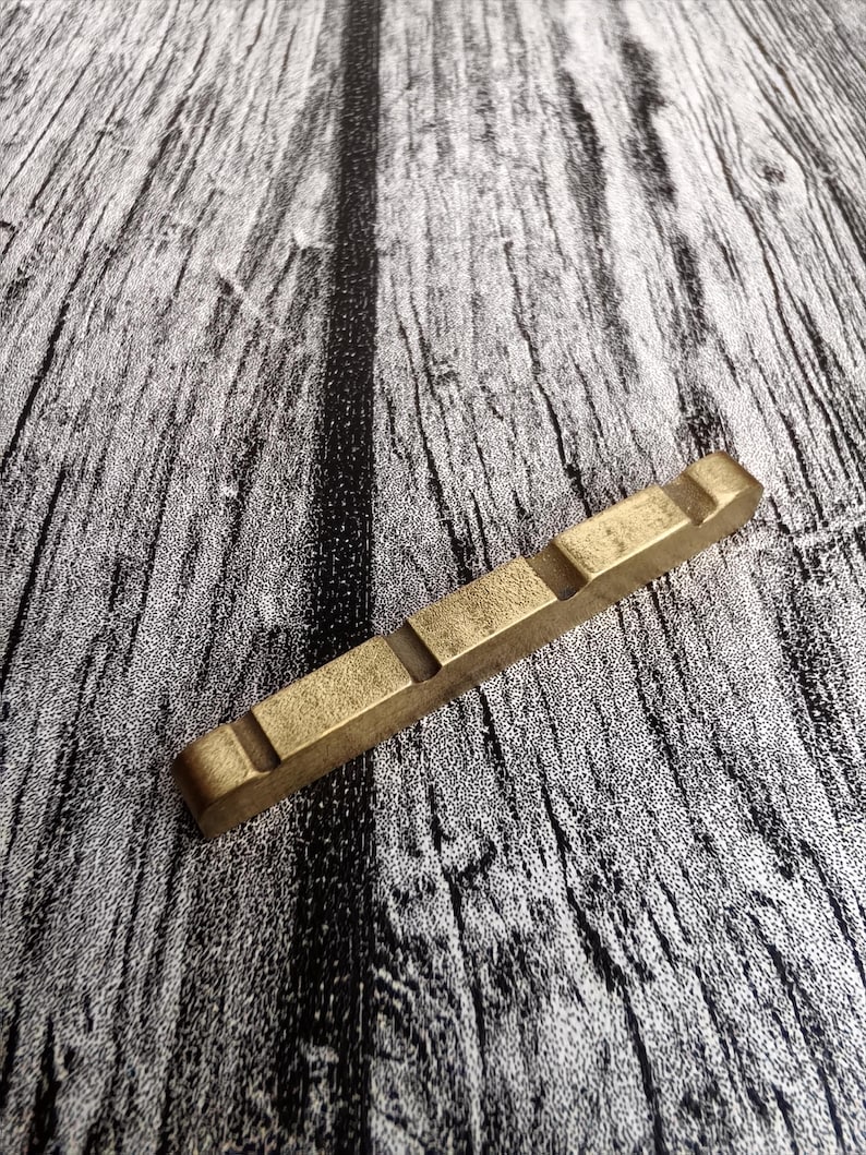 May include: A rectangular, brass-colored guitar nut with four grooves. The nut is made of a solid, metallic material and is designed to fit on a guitar. The background is a gray and white wood grain pattern.