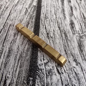 May include: A rectangular, brass-colored metal object with a series of recessed sections. The object is lying on a textured, gray and black wooden surface. The metal has a matte finish.