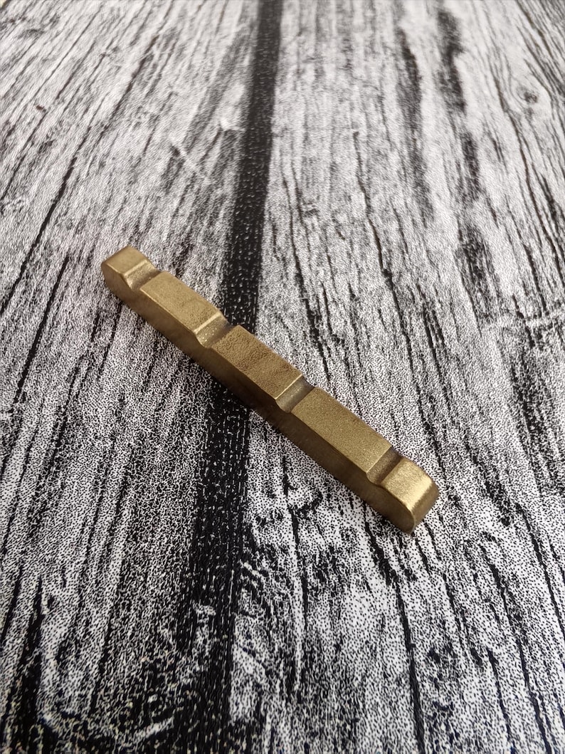 May include: A rectangular, brass-colored metal object with a series of recessed sections. The object is lying on a textured, gray and black wooden surface. The metal has a matte finish.