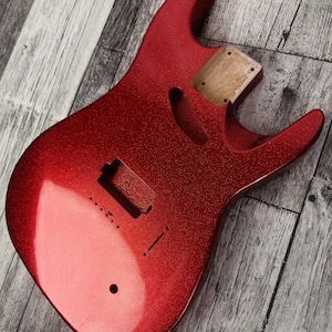 May include: A red electric guitar body with a sparkly finish. The body has cutouts for pickups, a tremolo system, and a neck. The wood grain is visible in the neck pocket. The guitar body is shown on a gray wooden surface.