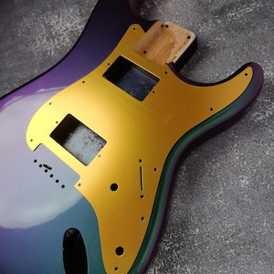 May include: A purple, green, and gold electric guitar body. The body is made of wood and has a gold pickguard.