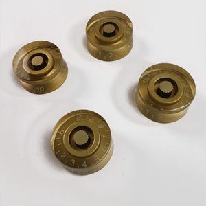 Könnte beinhalten: Vier goldfarbene Gitarrenknöpfe mit weißen Zahlen und Markierungen. Die Knöpfe sind rund und haben eine erhabene Mitte.