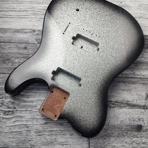 Könnte beinhalten: Ein silber-schwarzer E-Gitarrenkorpus mit glitzerndem Finish. Der Korpus hat Aussparungen für Tonabnehmer und einen Hals, und ein Holzstück ist befestigt. Der Gitarrenkorpus liegt auf einer Holzoberfläche.