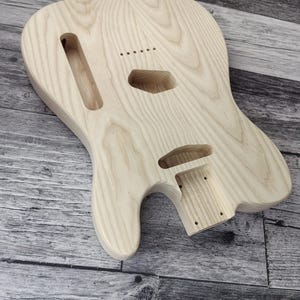 Pode incluir: Um corpo de guitarra elétrica de madeira clara sem acabamento com um padrão de grão visível. O corpo apresenta cavidades fresadas para captadores e eletrônicos, um encaixe para o braço e orifícios para as cordas. Texto Havok e Made in Canada.