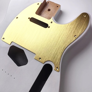 May include: A white guitar body with a gold-coloured metal pickguard and control plate. The pickguard has a brushed metal texture and is secured with screws. The guitar body has cutouts for pickups and controls, and a wooden neck pocket.