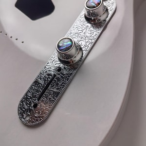 Puede incluir: Primer plano de una placa de control de guitarra cromada con dos perillas. La placa tiene un diseño floral ornamentado. Las perillas tienen una incrustación de nácar. El cuerpo de la guitarra es blanco.