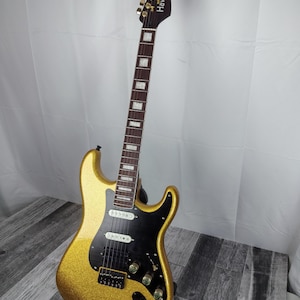 以下が含まれることがあります： 黒いピックガードと金色のハードウェアを備えた、金色のグリッターのエレキギター。ギターは、ブランド名「Havok」が白で書かれた濃い茶色のヘッドストックを持っています。指板には長方形のインレイがあります。ギターは黒いスタンドに置かれています。