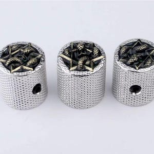 Puede incluir: Tres pomos de guitarra plateados con un diseño de estrella negra y dorada. Cada pomo tiene una superficie texturizada y un pequeño agujero en el centro.