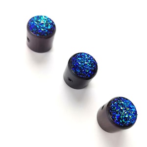 May include: Three dark brown cylindrical objects with a sparkling blue top. The tops of the cylinders are covered in a glittery, textured material. The objects are evenly spaced against a white background.