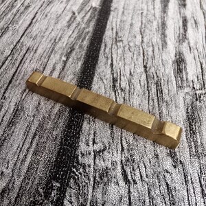 May include: A rectangular, brass-colored metal object with a series of rectangular cutouts. The object is lying on a textured, gray and black wooden surface. The metal piece has a slightly worn appearance.