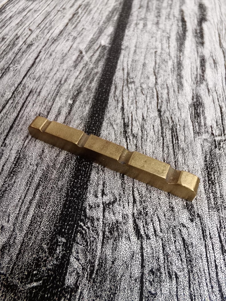 May include: A rectangular, brass-colored metal object with a series of rectangular cutouts. The object is lying on a textured, gray and black wooden surface. The metal piece has a slightly worn appearance.