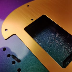 May include: A gold-colored metal guitar pickguard with a cutout for a pickup. The pickguard is on a guitar body with a green, blue, and purple iridescent finish.