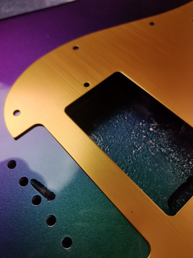 May include: A gold-colored metal guitar pickguard with a cutout for a pickup. The pickguard is on a guitar body with a green, blue, and purple iridescent finish.