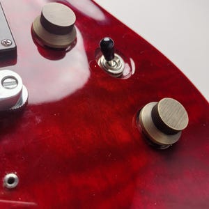 Może przedstawiać: Zbliżenie na czerwony korpus gitary elektrycznej z mosiężnymi pokrętłami i czarnym przełącznikiem dźwigienkowym. Wykończenie gitary jest błyszczące, z widocznym usłojeniem drewna. Pokrętła są okrągłe i teksturowane.