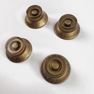 Könnte beinhalten: Vier goldfarbene Gitarrenknöpfe mit weißen Zahlen und Markierungen. Die Knöpfe sind rund mit einer erhabenen Mitte und einer flachen Basis.