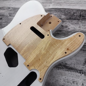 May include: A white electric guitar body with a natural wood pickguard and control plate. The guitar body has a smooth, glossy finish. The pickguard and control plate are made of light-coloured wood with visible grain. The guitar is partially assembled.