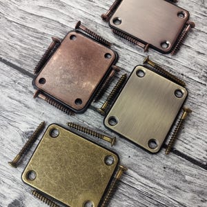 May include: Four rectangular metal neck plates for guitars, each featuring four corner screw holes and accompanied by four matching screws. The plates are finished in distinct metallic tones including brushed copper, aged brass, and polished nickel.