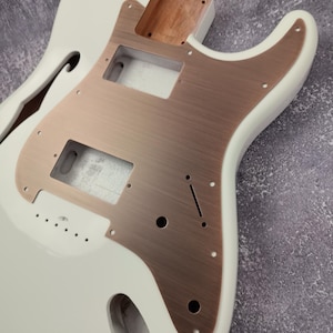 Puede incluir: Cuerpo de guitarra eléctrica blanco con una golpeador de color cobre. El cuerpo de la guitarra tiene un diseño de corte y está listo para ensamblar.