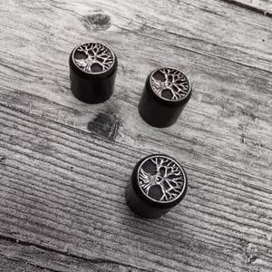 Könnte beinhalten: Drei schwarze Ohrstecker mit einem silbernen Lebensbaum-Design. Die Ohrstecker sind zylindrisch mit einer flachen Oberseite, die die detaillierte Baumgrafik zeigt. Der Hintergrund ist eine graue Holzoberfläche.