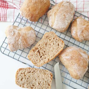 May include: A close-up of a cooling rack with six freshly baked bread rolls. The rolls are golden brown and have a slightly crispy crust. A silver knife is placed on the rack.