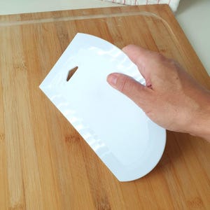 May include: A white plastic scraper with a rounded edge and a hole in the handle. The scraper is on a wooden cutting board.