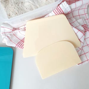 May include: Two beige plastic dough scrapers, one larger than the other, on a white surface with a red and white checkered kitchen towel.