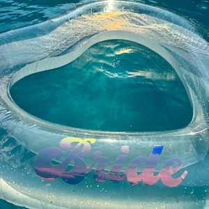 May include: A clear inflatable heart-shaped pool float with the word "Bride" in iridescent pink, blue, and yellow letters. The float is in a blue swimming pool.