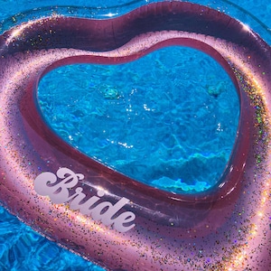 Puede incluir: Un flotador de piscina rosa en forma de corazón con detalles de purpurina y la palabra "Bride" en escritura blanca. El flotador está en una piscina de agua azul, creando un ambiente veraniego y festivo. Perfecto para una despedida de soltera o una fiesta de compromiso.