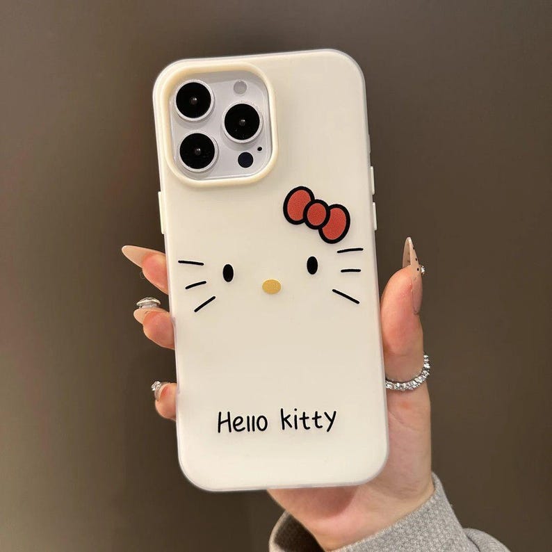 Puede incluir: Una funda blanca para tel&eacute;fono con un dise&ntilde;o de Hello Kitty. La funda tiene un lazo rojo, bigotes negros y una nariz amarilla. Las palabras "Hello Kitty" est&aacute;n impresas en negro en la parte inferior de la funda.