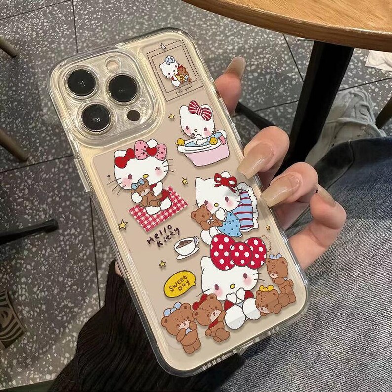 Peut inclure: Coque de t&eacute;l&eacute;phone transparente orn&eacute;e de motifs Hello Kitty, d'ours en peluche et de n&oelig;uds, avec les inscriptions "Hello Kitty" et "Sweet Day". Le bo&icirc;tier a un fond beige et est tenu par une personne.