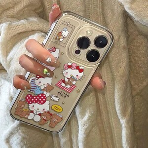 Peut inclure: Coque de t&eacute;l&eacute;phone transparente orn&eacute;e de motifs Hello Kitty et ours en peluche. L'&eacute;tui a un fond beige avec diverses images de Hello Kitty avec des n&oelig;uds rouges et des ours en peluche. L'&eacute;tui a une bordure transparente et recouvre l'appareil photo du t&eacute;l&eacute;phone.