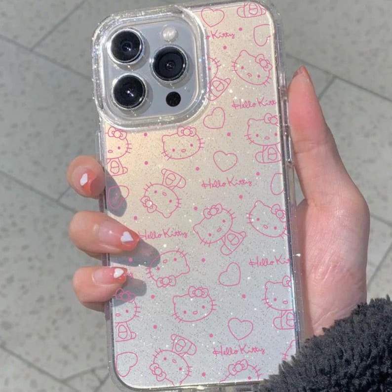 K&ouml;nnte beinhalten: Transparente Handyh&uuml;lle mit einem rosa Hello Kitty Muster und Glitzerakzenten. Die H&uuml;lle zeigt mehrere Hello Kitty Illustrationen, Herzen und den Text "Hello Kitty". Die H&uuml;lle ist f&uuml;r ein Smartphone konzipiert.