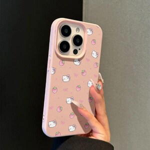 Może przedstawiać: R&oacute;żowe etui na telefon z powtarzającym się wzorem twarzy Hello Kitty, truskawek i kokardek. Etui ma podwyższoną krawędź wok&oacute;ł obiektyw&oacute;w aparatu i gładkie wykończenie. Zaprojektowane dla smartfona.