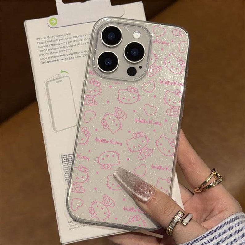 K&ouml;nnte beinhalten: Transparente iPhone 15 Pro H&uuml;lle mit einem rosa Hello Kitty Muster. Die H&uuml;lle hat ein glitzerndes Finish und zeigt mehrere Hello Kitty Illustrationen und Herzen. Die Handyh&uuml;lle wird in einer Hand mit Ringen gehalten.