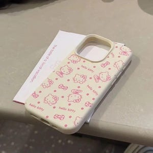May include: A cream-coloured phone case with a repeating pattern of pink Hello Kitty characters and bows. The case has a protective design with cutouts for the camera and buttons. The text "hello kitty" is printed on the case.