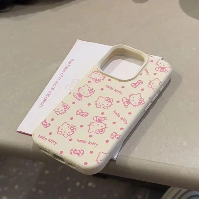 May include: A cream-coloured phone case with a repeating pattern of pink Hello Kitty characters and bows. The case has a protective design with cutouts for the camera and buttons. The text "hello kitty" is printed on the case.