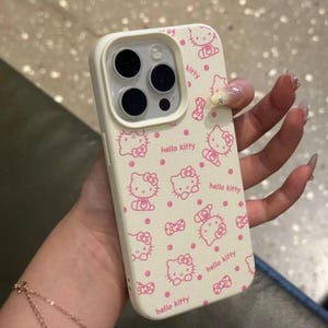 May include: Cream-coloured phone case with a pink Hello Kitty design. The pattern includes Hello Kitty characters, bows, and polka dots. The case has a camera cutout and is held in a hand.