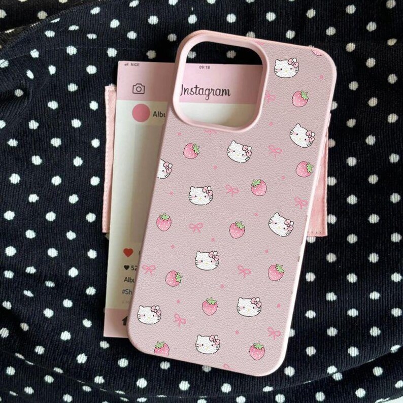 Może przedstawiać: R&oacute;żowe etui na telefon z motywami Hello Kitty, truskawek i kokardek. Etui ma wycięcie na aparat. R&oacute;żowa karta z motywem Instagrama znajduje się za etui. Czarne tło z białymi kropkami.
